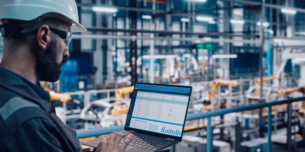 Manufacturing setting. A map wearing a helmet and protective eyewear, typing on a laptop.